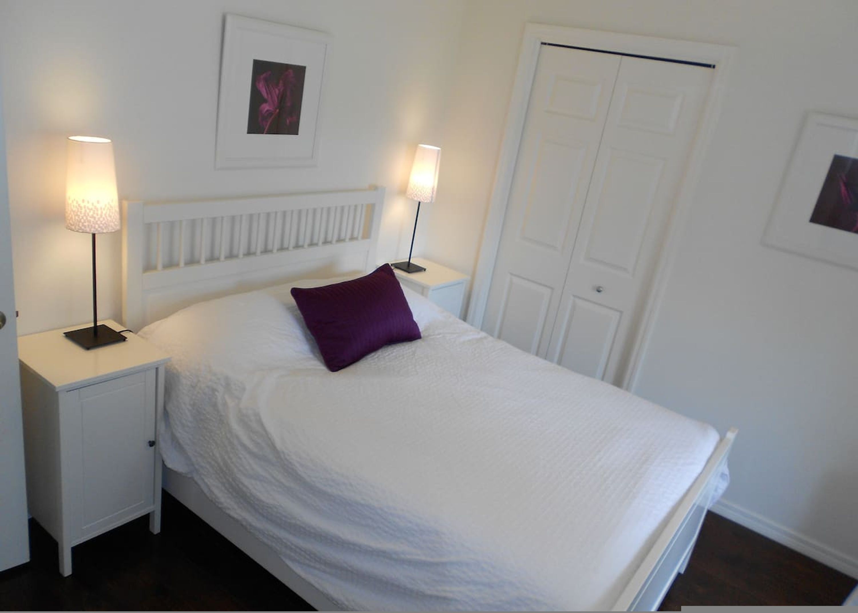 A neatly made bed with a white comforter and a purple pillow, two bedside lamps, a white radiator, and a white door.