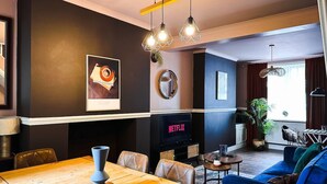 A modern living room with a wooden dining table, leather chairs, a blue sofa, a TV, and decorative wall art.
