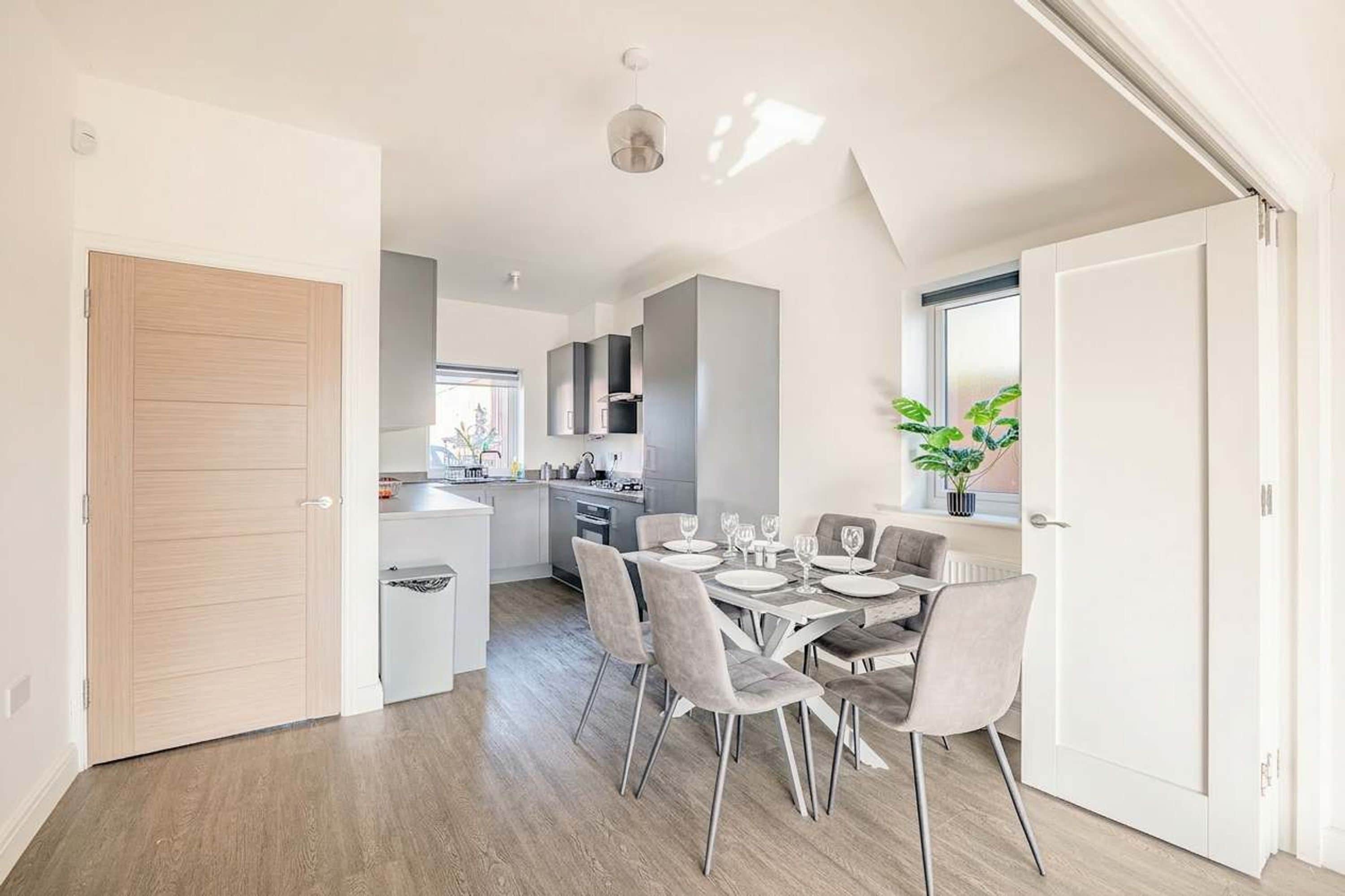 A modern kitchen with a central island, a dining area with grey chairs, and a large window with a plant.