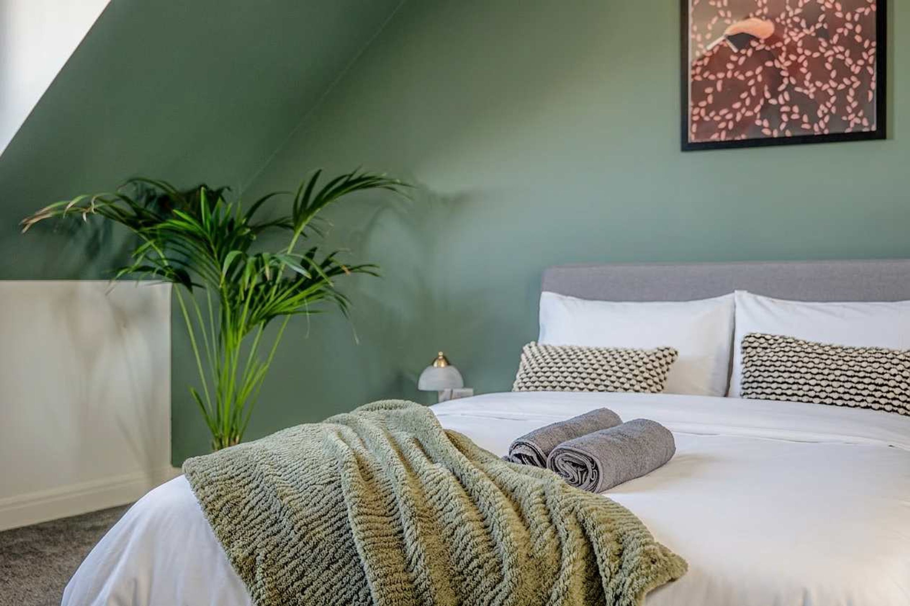 Une chambre à coucher avec un lit, des oreillers, une couverture, une plante et une œuvre d’art murale.