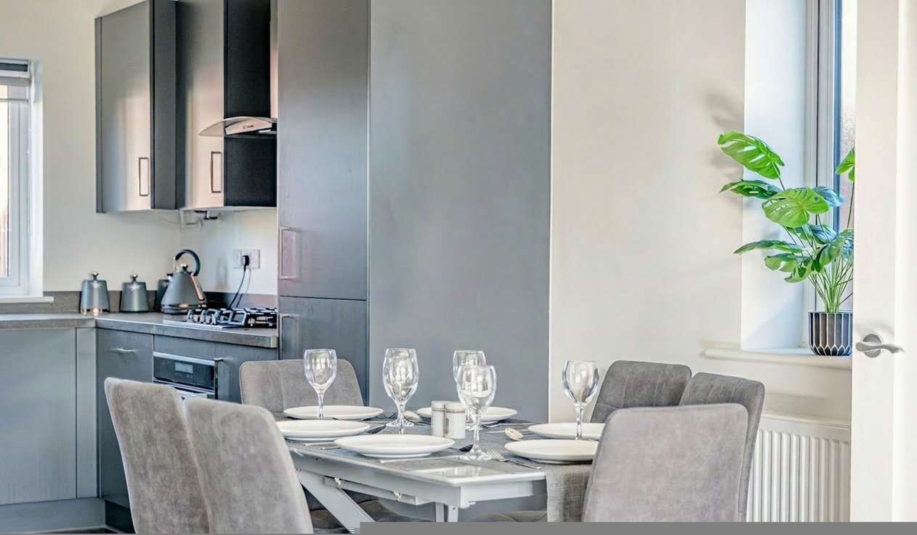 A modern kitchen with grey cabinets, a dining table set for four, and a potted plant by the window.