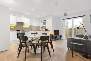 A modern kitchen with white cabinets, a dining table with chairs, and a grey sofa in a bright living area with a large window. - Garden Flat in Central London (London)