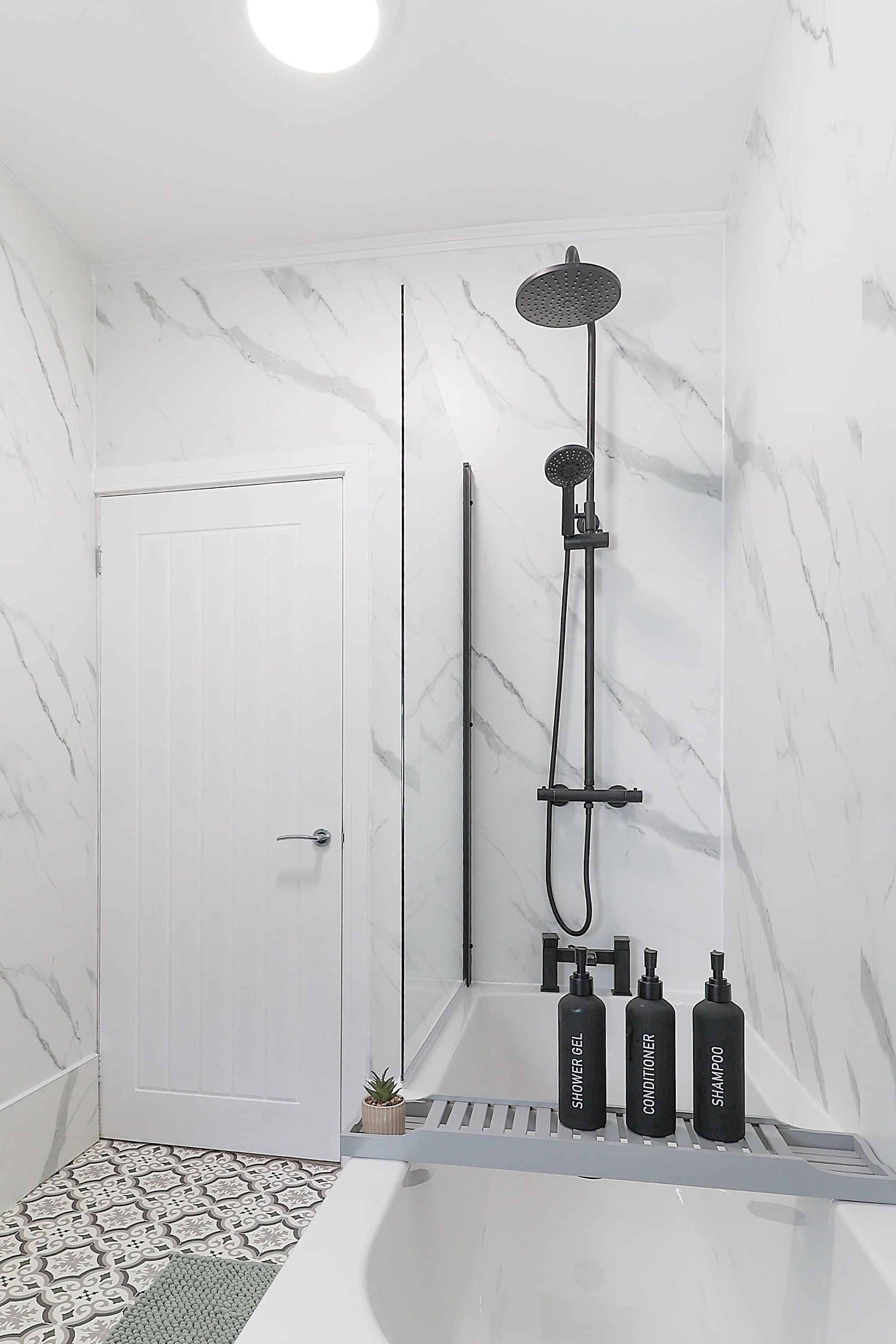 A bathroom with a white door, a shower with a handheld showerhead, and three bottles labeled 'shower gel,' 'conditioner,' and 'shampoo' on a shelf.