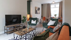 A living room with a TV, two green armchairs, a sofa, a coffee table, and a dining area in the background. - Perfect Booking 4 Bed Home in Aberdare (Aberdare)