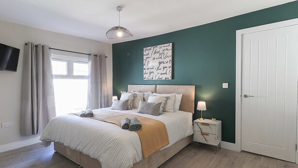 A bedroom with a bed, a bedside table with a lamp, a wall-mounted TV, and a door. - Perfect Booking 4 Bed Home in Aberdare (Aberdare)