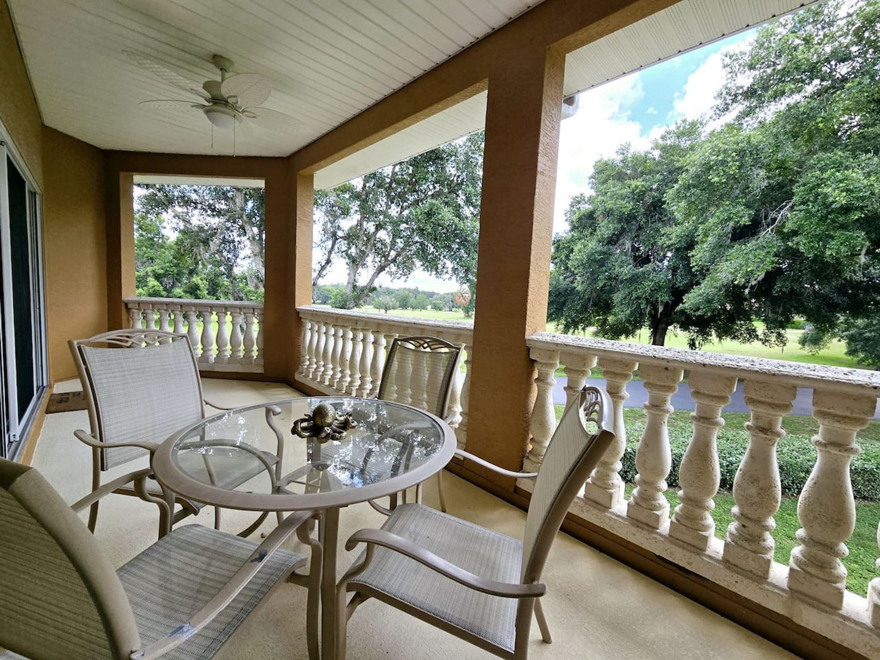A covered outdoor seating area with a glass table and chairs, a ceiling fan, and a view of a landscaped garden.