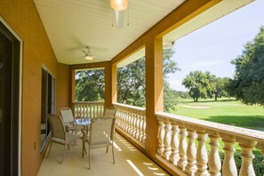 A covered balcony with a table and chairs, a view of a golf course, and a ceiling fan.