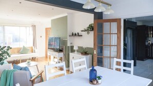 A modern living room with a sofa, dining table, and chairs. There is a television, a small table with decorative items, and a kitchen area visible through an open door. - Perfect Booking 4 Bed Home Near City Airport (Cheadle)