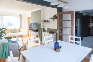 A modern living room with a sofa, dining table, and chairs. There is a television, a small table with decorative items, and a kitchen area visible through an open door.