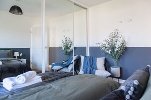 A modern hotel room with a bed, a chair, a mirror, and two potted plants.