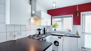 A modern kitchen with white cabinets, a black countertop, a built-in washing machine, and a window with a plant. - Perfect Booking 4 Bed Home in Leytonstone (London)