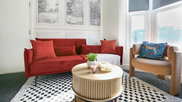 A room with a red sofa, a round coffee table with a plant, a blue cushion, and a checkered rug. - Perfect Booking 4 Bed Home in Leytonstone (London)