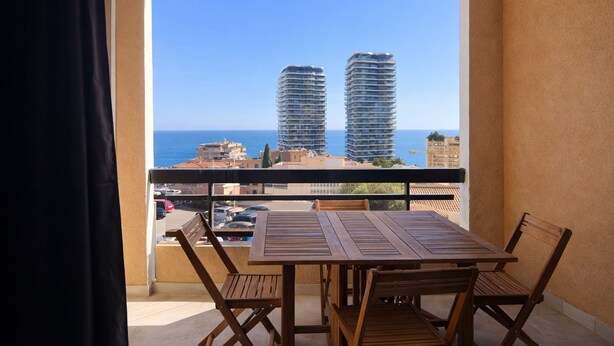 Un balcon avec une table et des chaises en bois, donnant sur un paysage urbain avec de hauts immeubles et un ciel bleu dégagé.