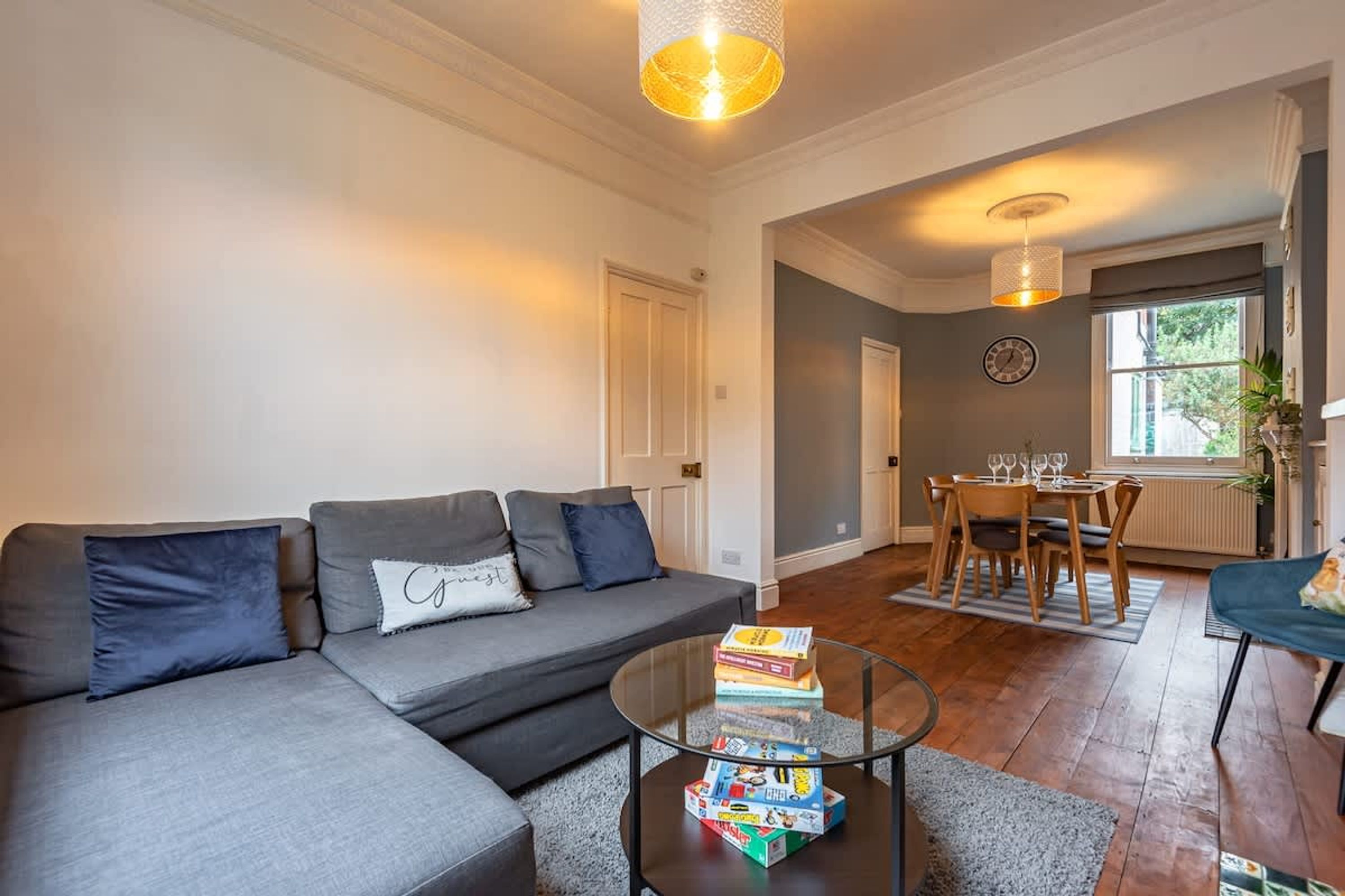 A living room with a sofa, a coffee table with books, a dining area with a table and chairs, and a window with a view.