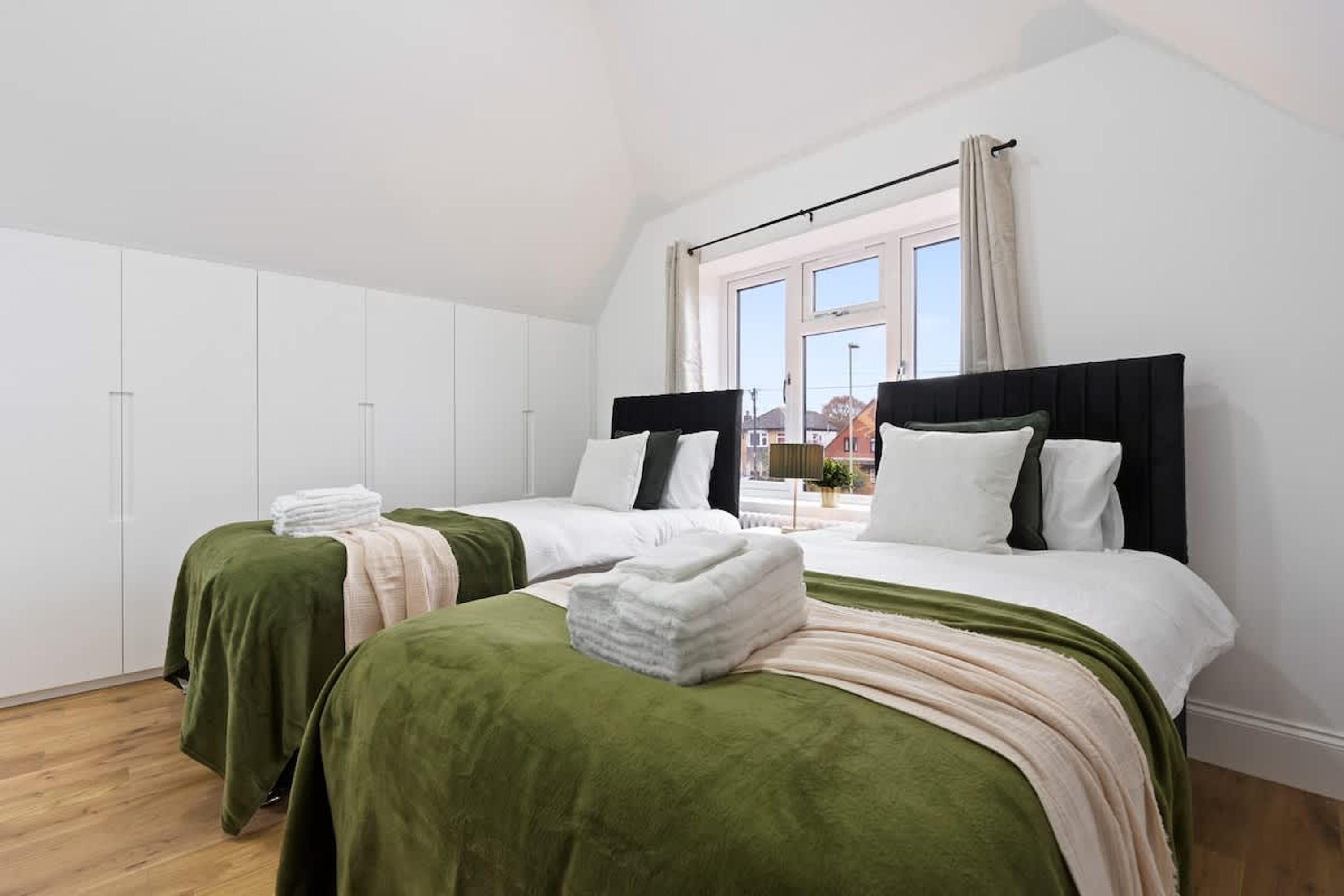 Een slaapkamer met twee bedden, witte muren, een raam met gordijnen en uitzicht op gebouwen.