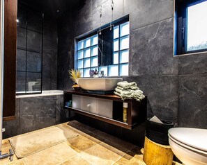 Um banheiro moderno com uma banheira grande, uma pia branca, uma janela com vários painéis e uma parede revestida de azulejos escuros.