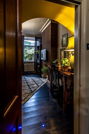 Ein Flur mit einer Holztreppe, einem Heizkörper, einem an der Wand befestigten Fernseher sowie einem Tisch mit Lampe und Pflanzen.