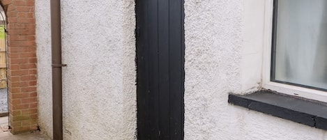 A white building with a black door and window, stone paving, and a brick wall.