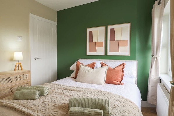 A bedroom with a bed, pillows, a bedside table with a lamp, and two framed artworks on the wall.