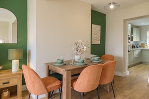 A dining area with a wooden table set for four, surrounded by orange chairs. A vase with flowers is on the table, and a lamp is on a side table.