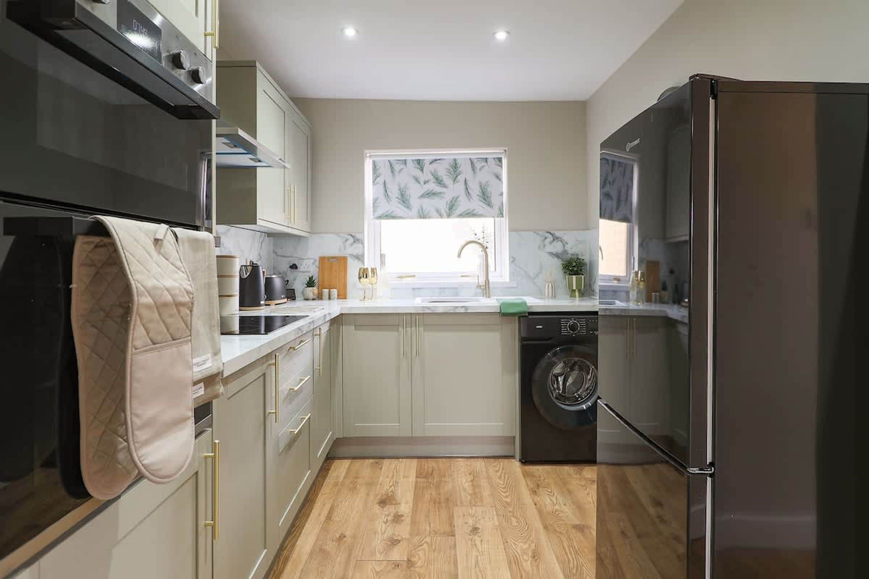 A compact kitchen with a washing machine, a refrigerator, and a window with a leaf-patterned blind.