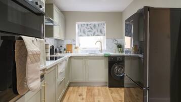 A compact kitchen with a washing machine, a refrigerator, and a window with a leaf-patterned blind.