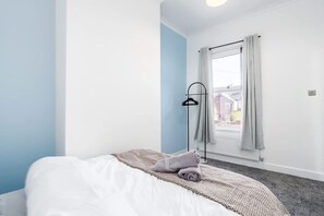 A hotel room with a bed, a window with curtains, a floor lamp, and a view of buildings outside. - Perfect Booking Stylish 3 Bed Family Home (Doncaster)