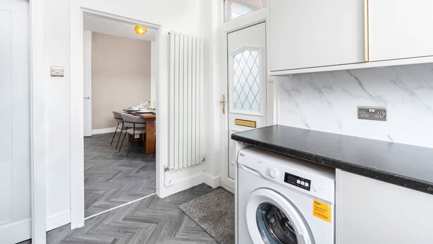 A compact laundry area with a washing machine, a radiator, and a dining area visible in the background. - Perfect Booking Stylish 3 Bed Family Home (Doncaster)