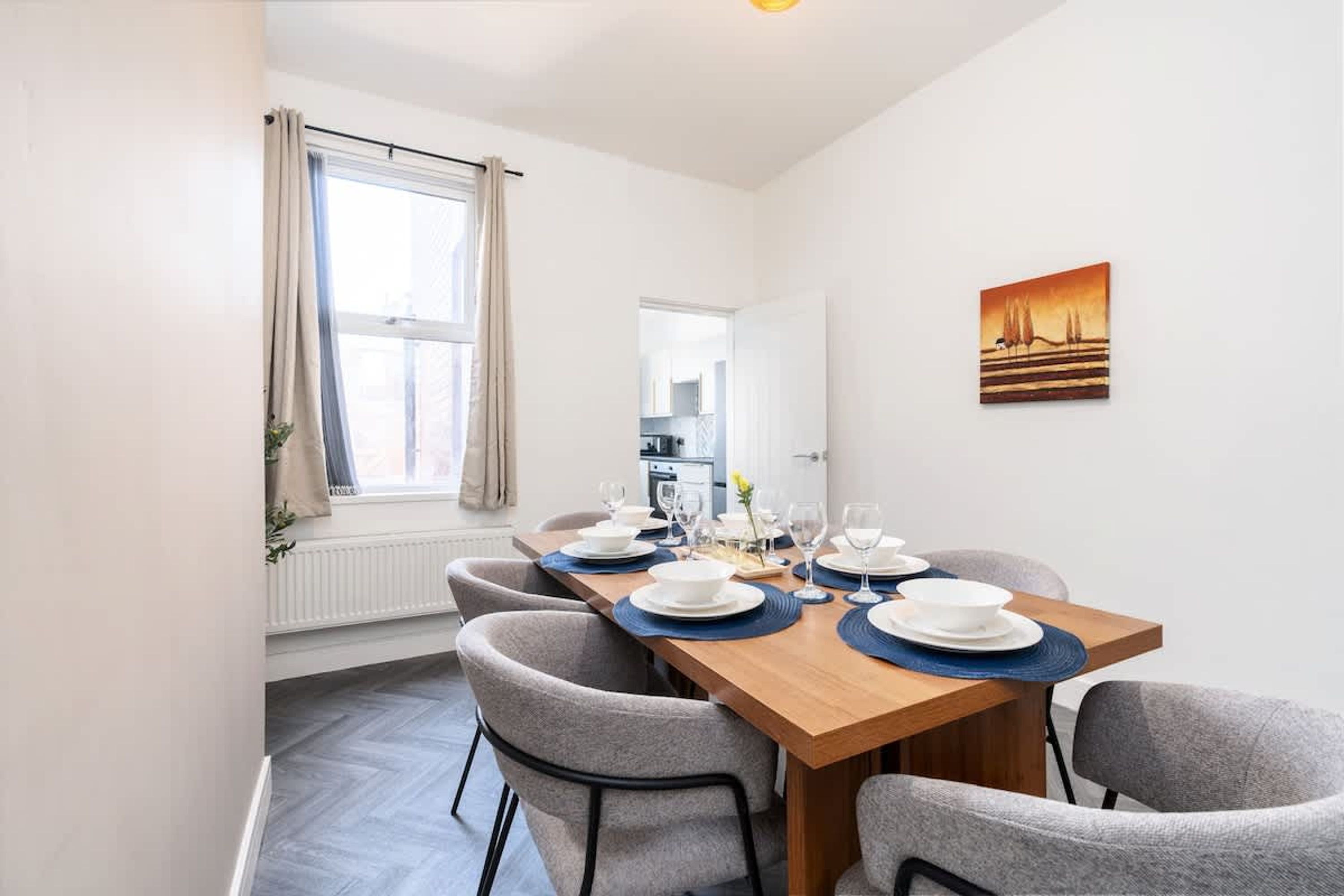 A dining area with a wooden table set for four, grey upholstered chairs, a window with curtains, and a painting on the wall.