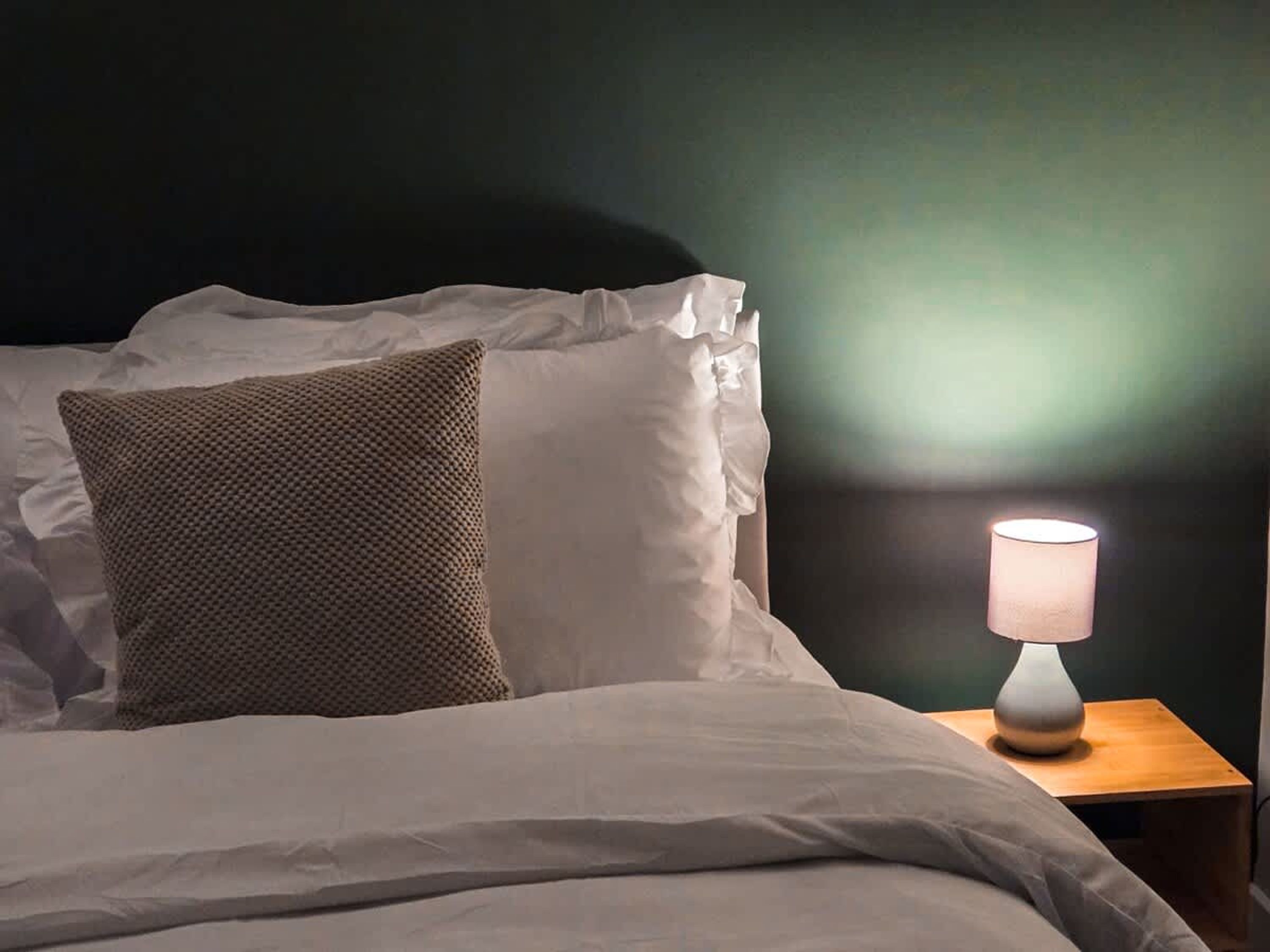 A bed with white bedding and a textured pillow, a bedside lamp, and a dark wall.