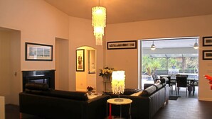 Ein Wohnzimmer mit Kamin, einer Kristallleuchter, einer Couch, einem Couchtisch und Blick durch eine Glastür auf einen Außenbereich.