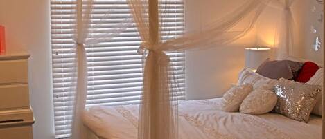 Ein Schlafzimmer mit einem Himmelbett, einem Nachttisch, einer Kommode und einem Fenster mit Jalousien.