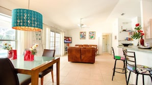 Uma sala de estar moderna com uma mesa de jantar de vidro, um sofá marrom, uma luminária pendente verde-água e uma área de cozinha com balcão e cadeiras.