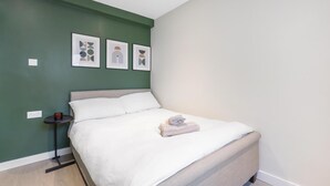 Ein ordentlich bezogenes Bett mit weißer Decke und einem gefalteten Handtuch, ein kleiner Nachttisch mit einem roten Gegenstand und drei gerahmte Kunstwerke an der grünen Wand.