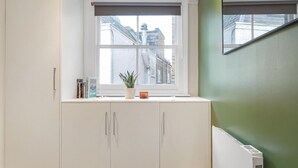 Eine Küche mit weißen Schränken, einem Fenster mit Blick auf Gebäude, einer kleinen Pflanze und einer Kerze auf dem Fensterbrett.