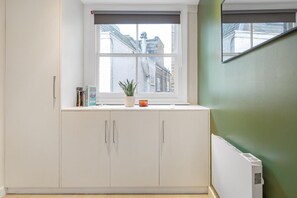 Eine Küche mit weißen Schränken, einem Fenster mit Blick auf Gebäude, einer kleinen Pflanze und einer Kerze auf dem Fensterbrett.