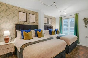 Hotelkamer met twee bedden, een nachtkastje, een lamp, ingelijste foto’s aan de muur en een raam met gordijnen.