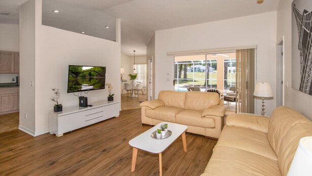 Uma sala de estar espaçosa com televisão de tela plana, um sofá bege, uma mesa de centro e uma área de jantar visível através de uma porta de vidro.