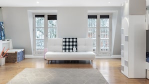 Una sala de estar moderna con un sofá blanco, dos ventanas con persianas, un tapete gris y un mueble blanco.