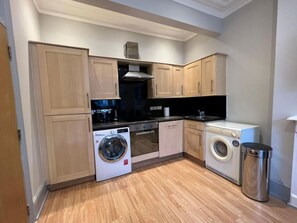 A compact kitchen with wooden cabinets, a washing machine, a dishwasher, and a trash bin. - Garden Flat in Central Battersea (London)
