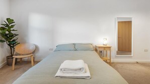 Un dormitorio con cama, mesita de noche con lámpara, una silla y una planta en maceta.