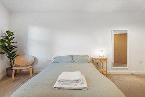 A bedroom with a bed, a bedside table with a lamp, a chair, and a potted plant. - Garden Flat in Central Battersea (London)