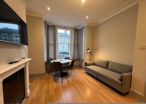 A living room with a sofa, a dining table with chairs, a TV mounted on the wall, and a window with curtains. - Garden Flat in Central Battersea (London)