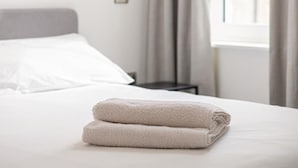 Una cama bien hecha con una toalla doblada, una ventana con cortinas y una mesita de noche pequeña.