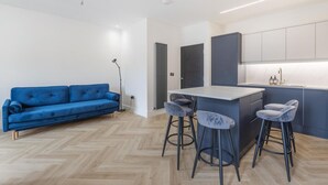 A modern kitchen with a bar area, a blue sofa, and two bar stools. - Modern Brixton Flat 2 (Magherafelt)