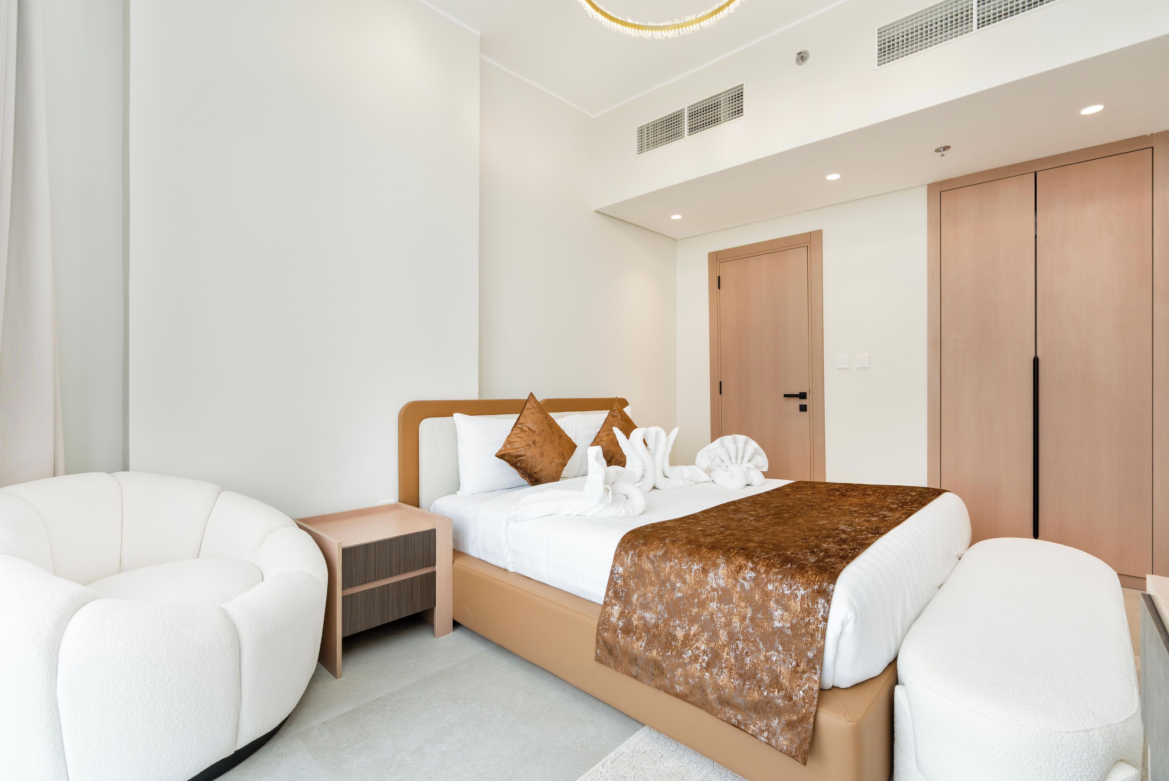 Une chambre d’hôtel moderne avec un grand lit, deux fauteuils blancs, une armoire en bois et une petite table de chevet.