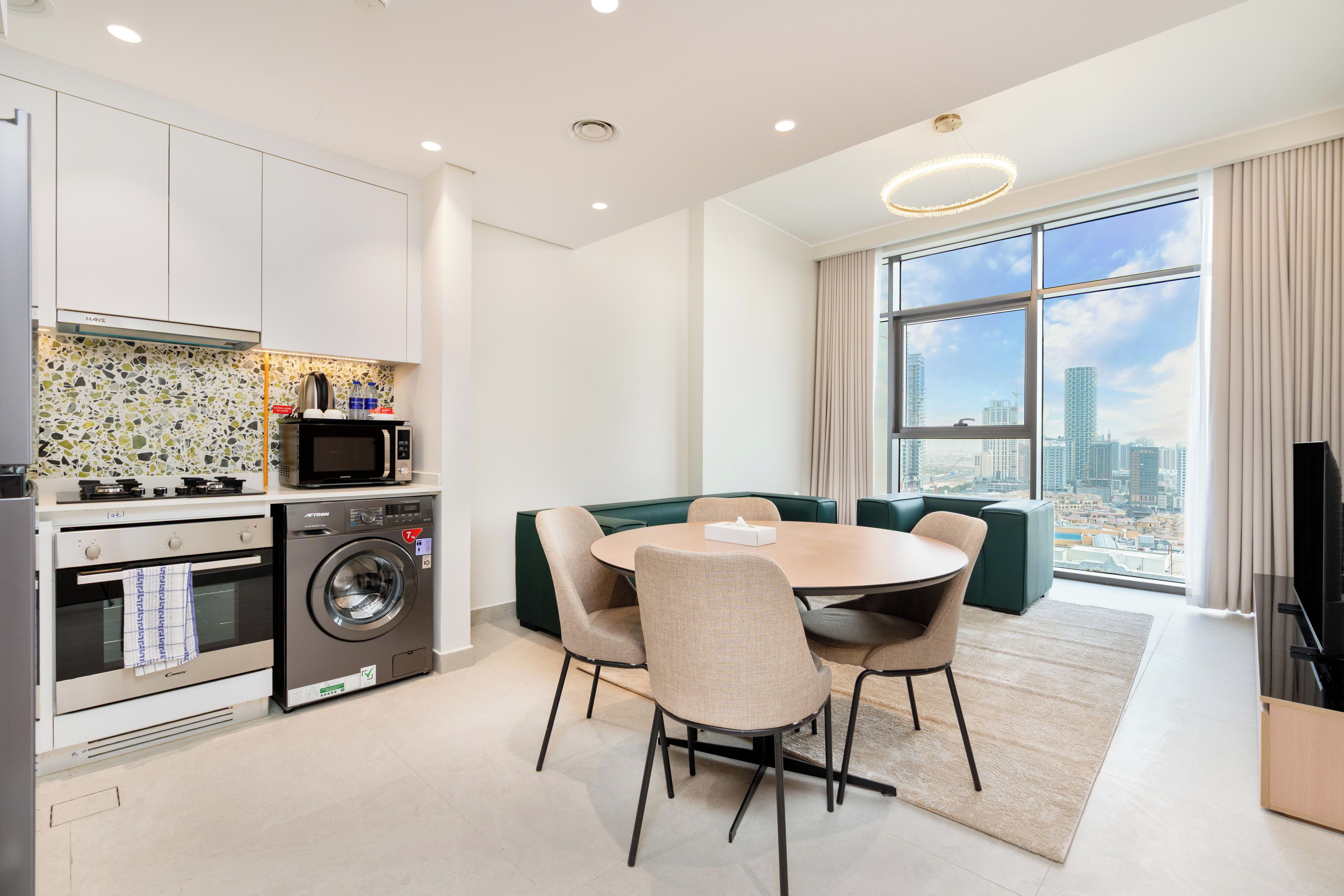 A modern kitchen with a built-in washer and dryer, a dining area with a round table and chairs, and a large window offering a city view.