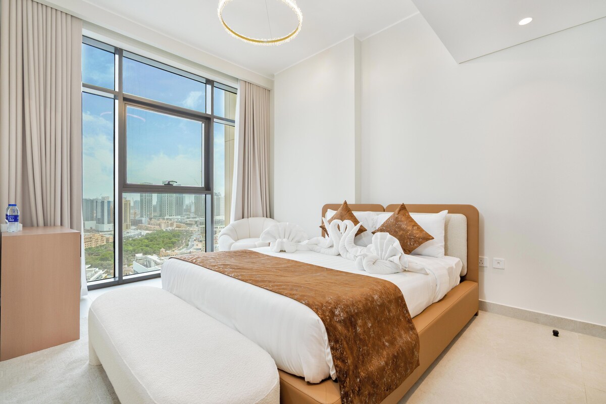 A hotel room with a large window, a bed with white bedding and brown accents, and a view of the cityscape.