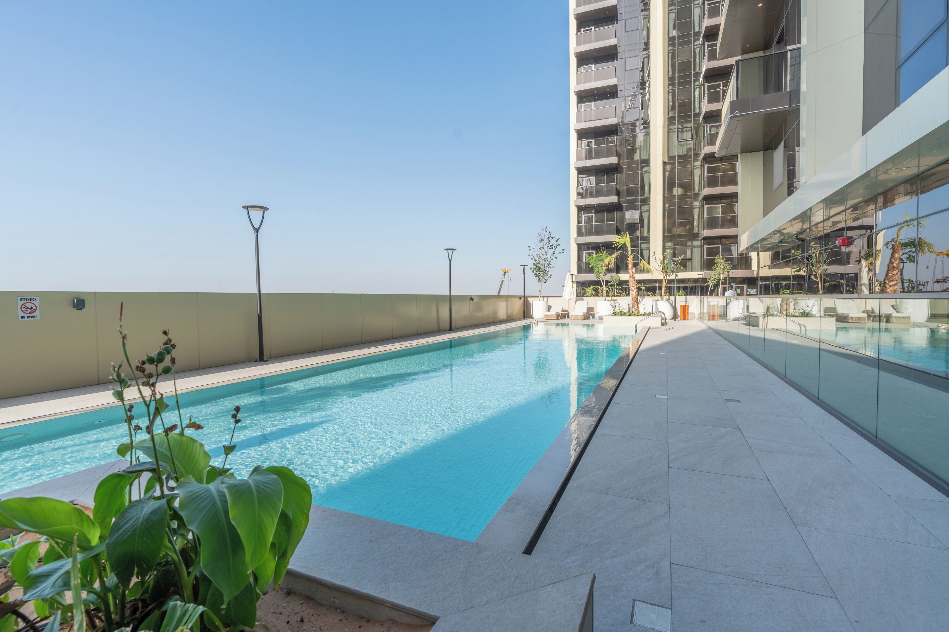 A swimming pool with a clear blue water, surrounded by a tiled walkway and some greenery.