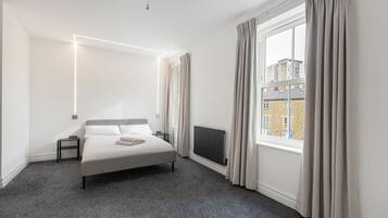 A modern bedroom with a large bed, a radiator, a window with curtains, and a view of buildings outside.